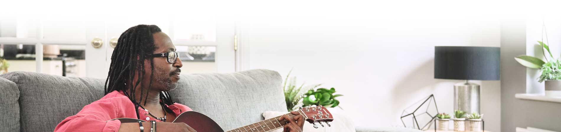 A person with dreadlocks plays guitar while sitting on a gray couch in a bright, modern living room.